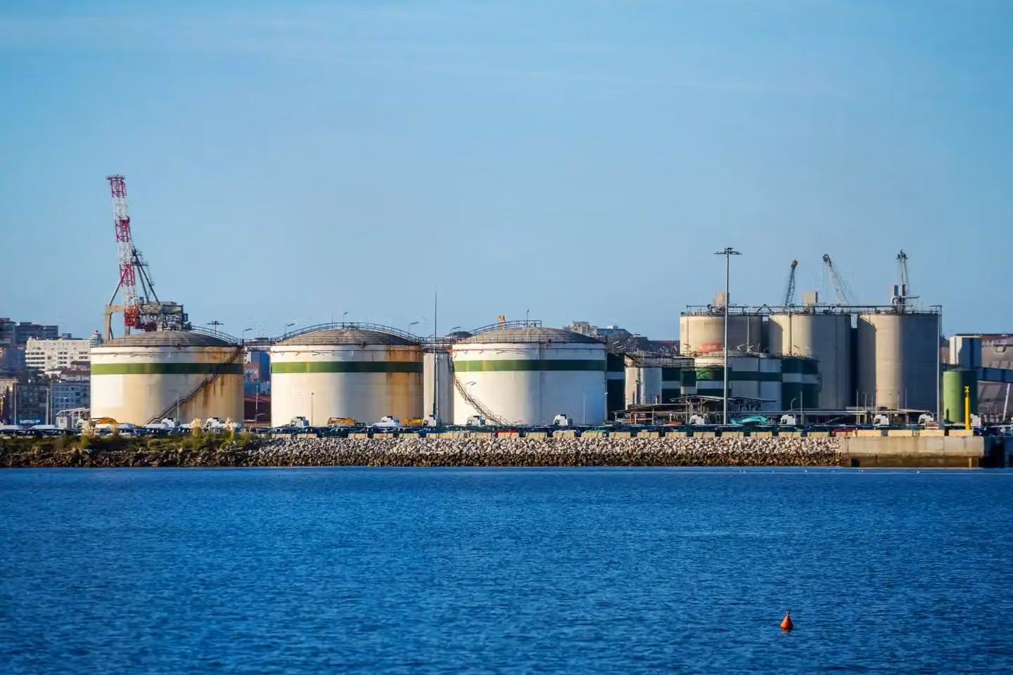 Large storage tanks at an industrial port facility by the water under clear blue skies.